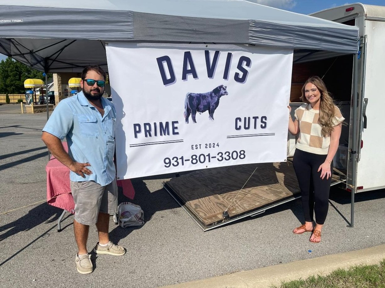 Two vendors stand in front of a food trailer displaying a sign for "Davis Prime Cuts," established in 2024. The sign features a cow graphic and includes a contact number, 931-801-3308, promoting fresh meat products. The setting appears to be a parking lot with sunny weather.