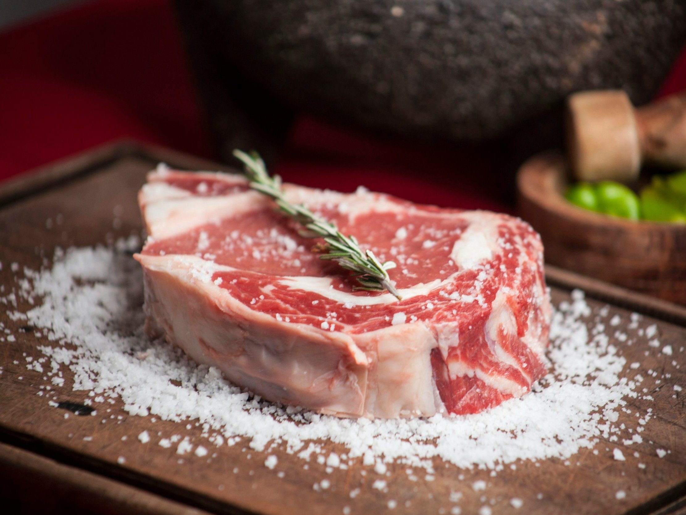 Freshly cut ribeye steak seasoned with salt and garnished with a sprig of rosemary, presented on a wooden cutting board, with a rustic background featuring a stone mortar and green vegetables.